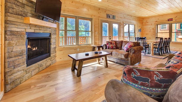 L’intérieur chaleureux d’un chalet avec une cheminée en pierre, un plancher en bois, un grand canapé et un ventilateur de plafond.