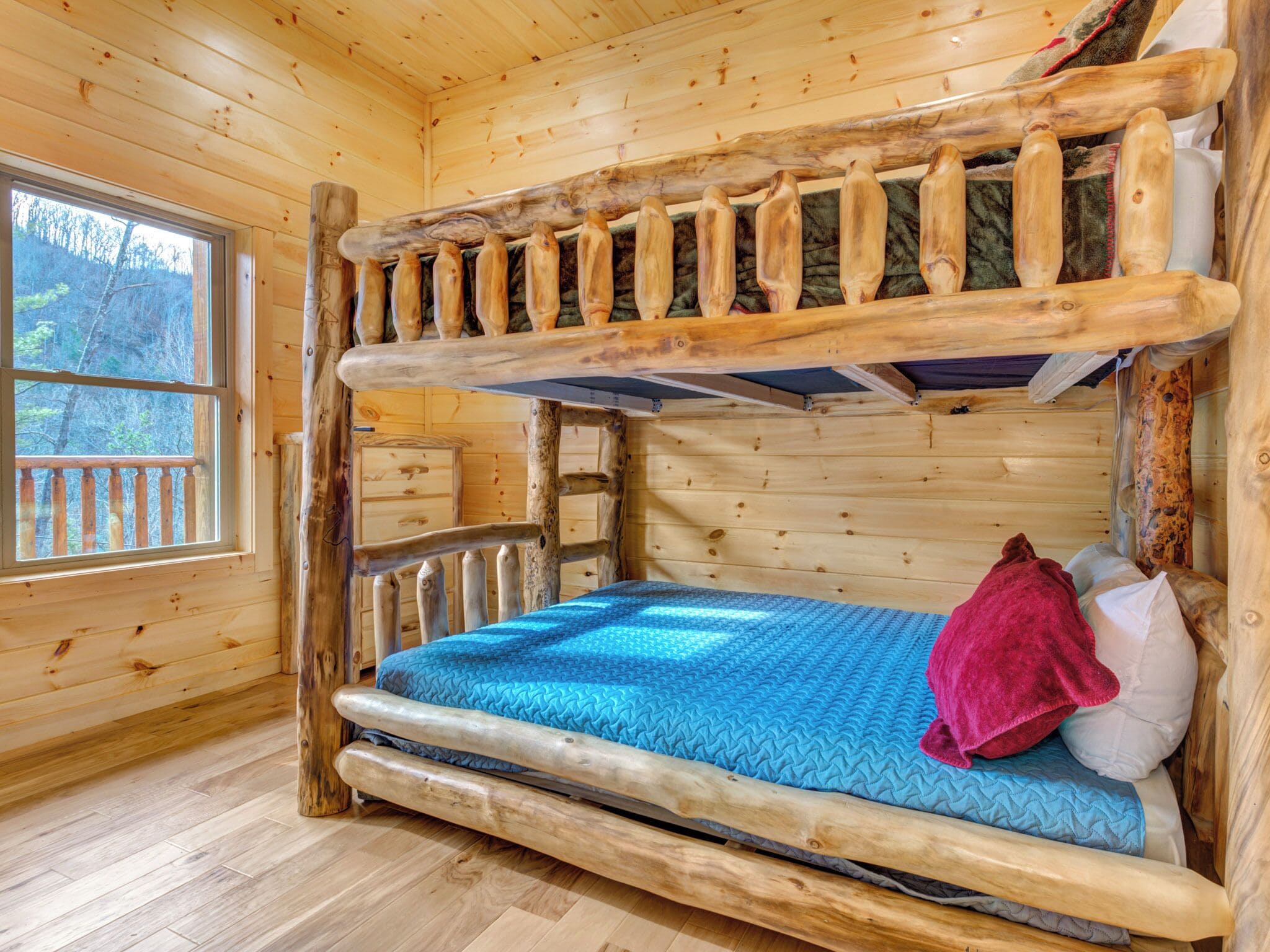 A wooden cabin room with a bunk bed, a window with a view of trees, and a wooden floor.