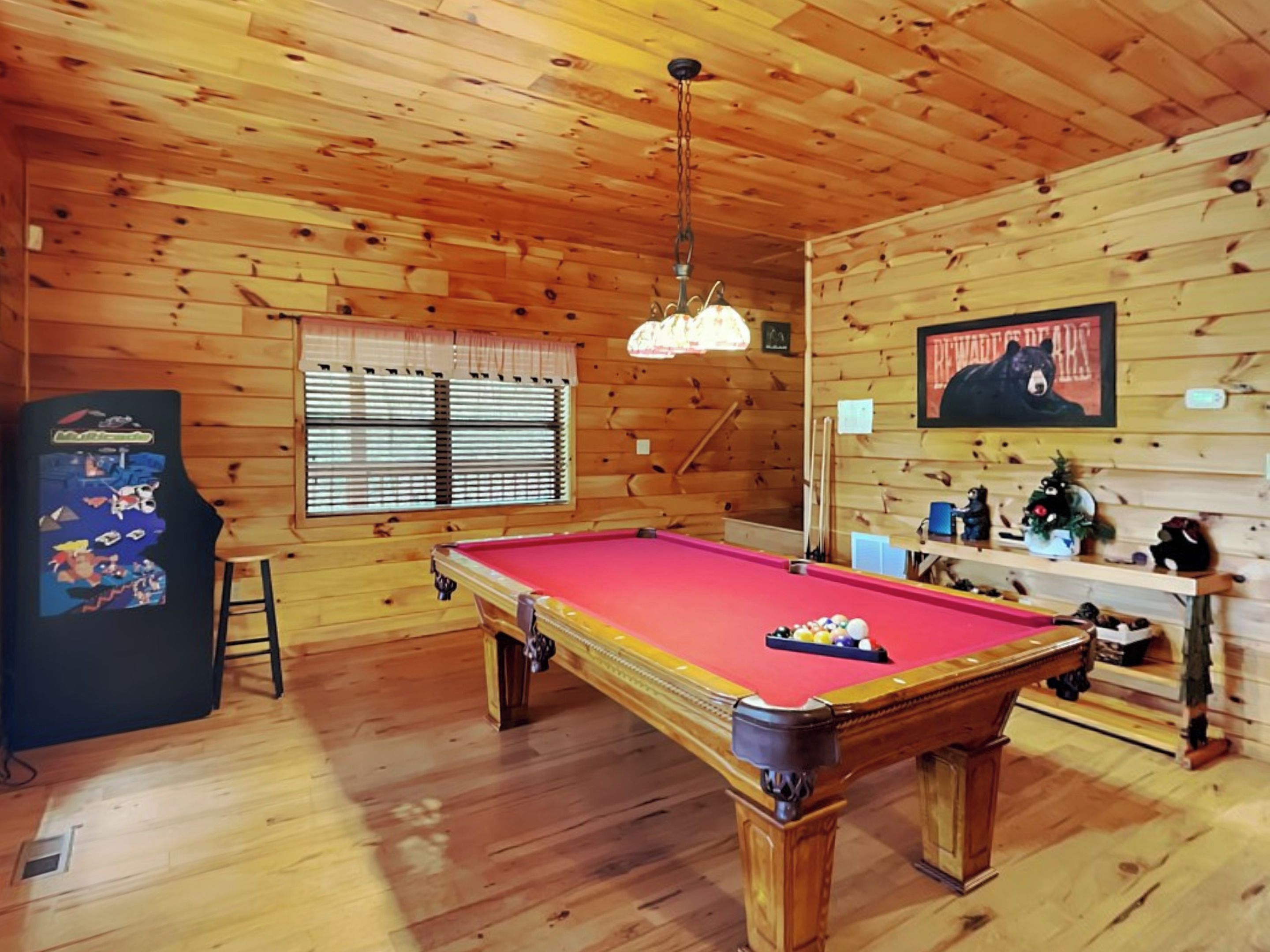 Une table de billard dans une pièce en bois, avec un ventilateur au plafond, un réfrigérateur et une affiche accrochée au mur.