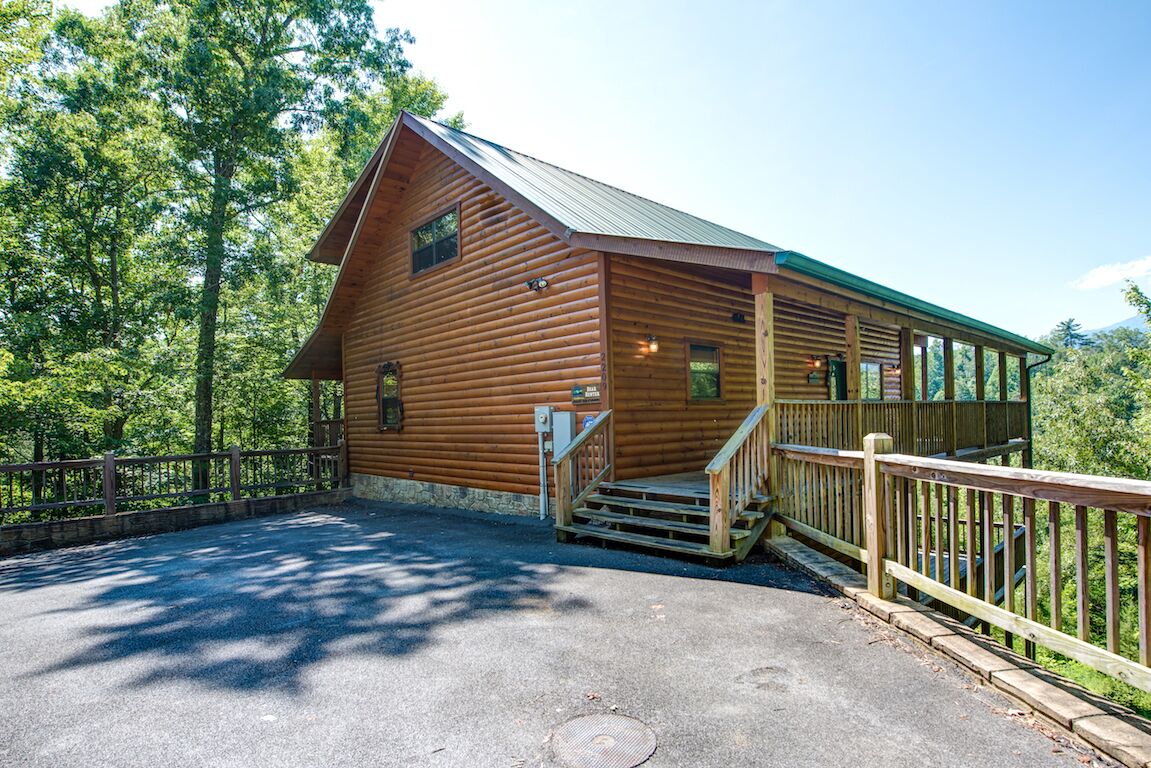 A wooden cabin with a metal roof, a staircase leading to a porch, and a fenced parking area.