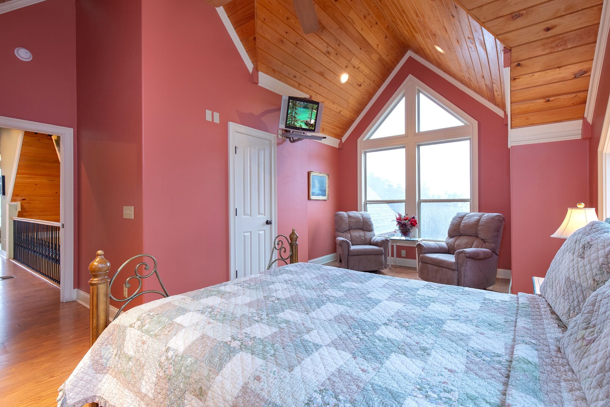 A bedroom with a large bed, two chairs, a TV mounted on the wall, and a window providing natural light.