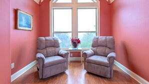 Um quarto com duas poltronas cinzentas, uma mesa pequena com um vaso de flores e uma janela grande com vista para árvores.