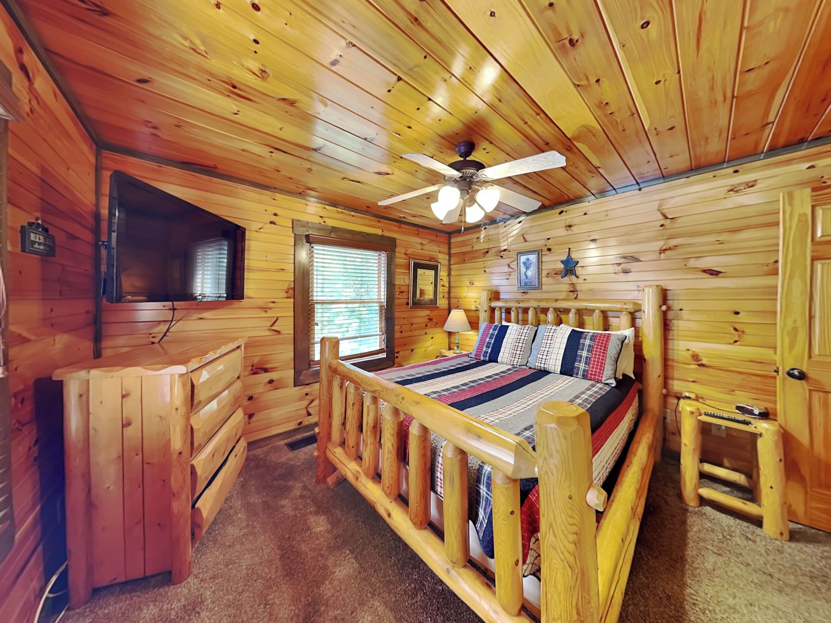 A wooden cabin room with a bed, a desk, a ceiling fan, and a window.