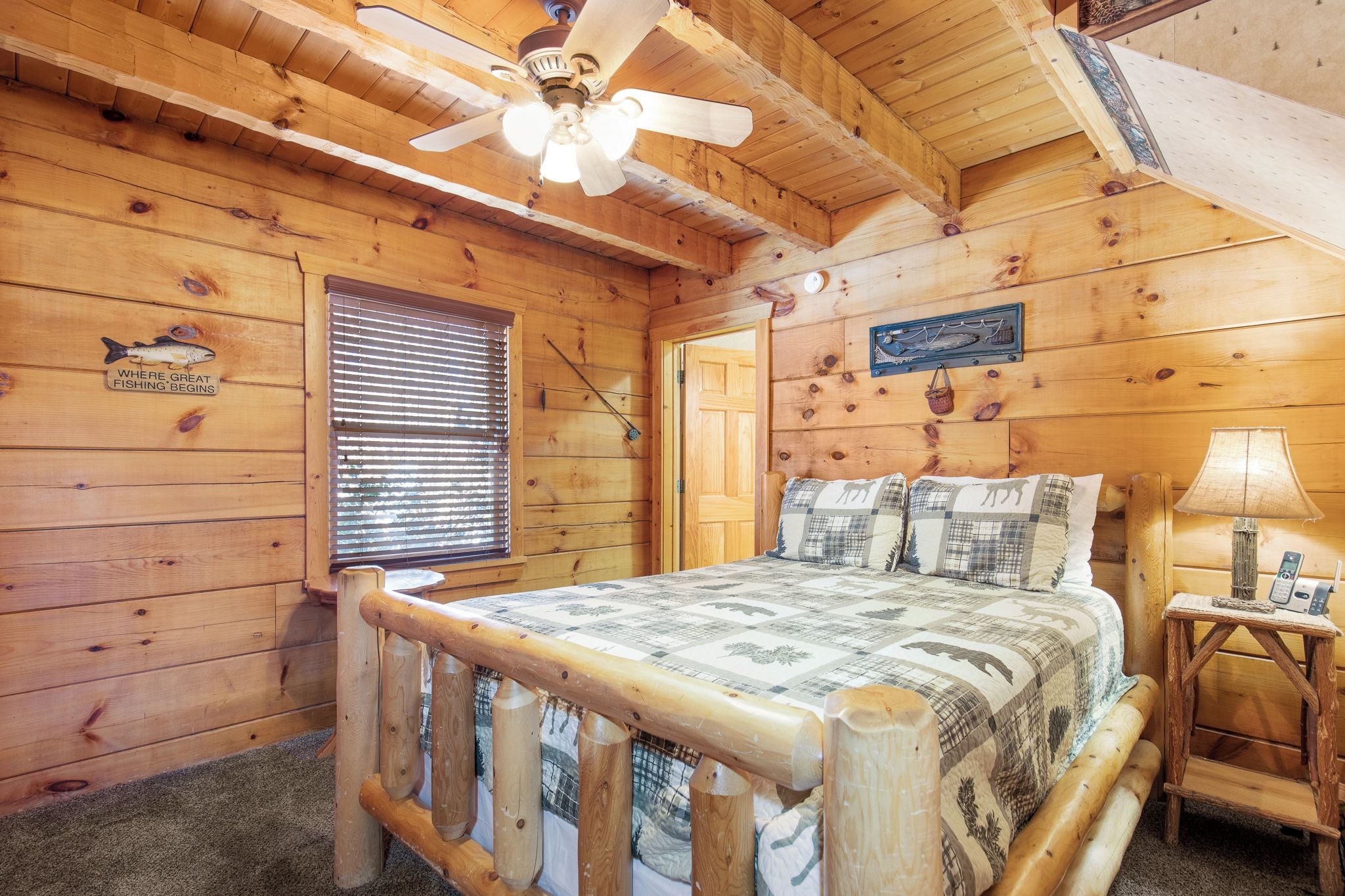 A wooden cabin bedroom with a bed, a ceiling fan, a window with blinds, and a sign on the wall.