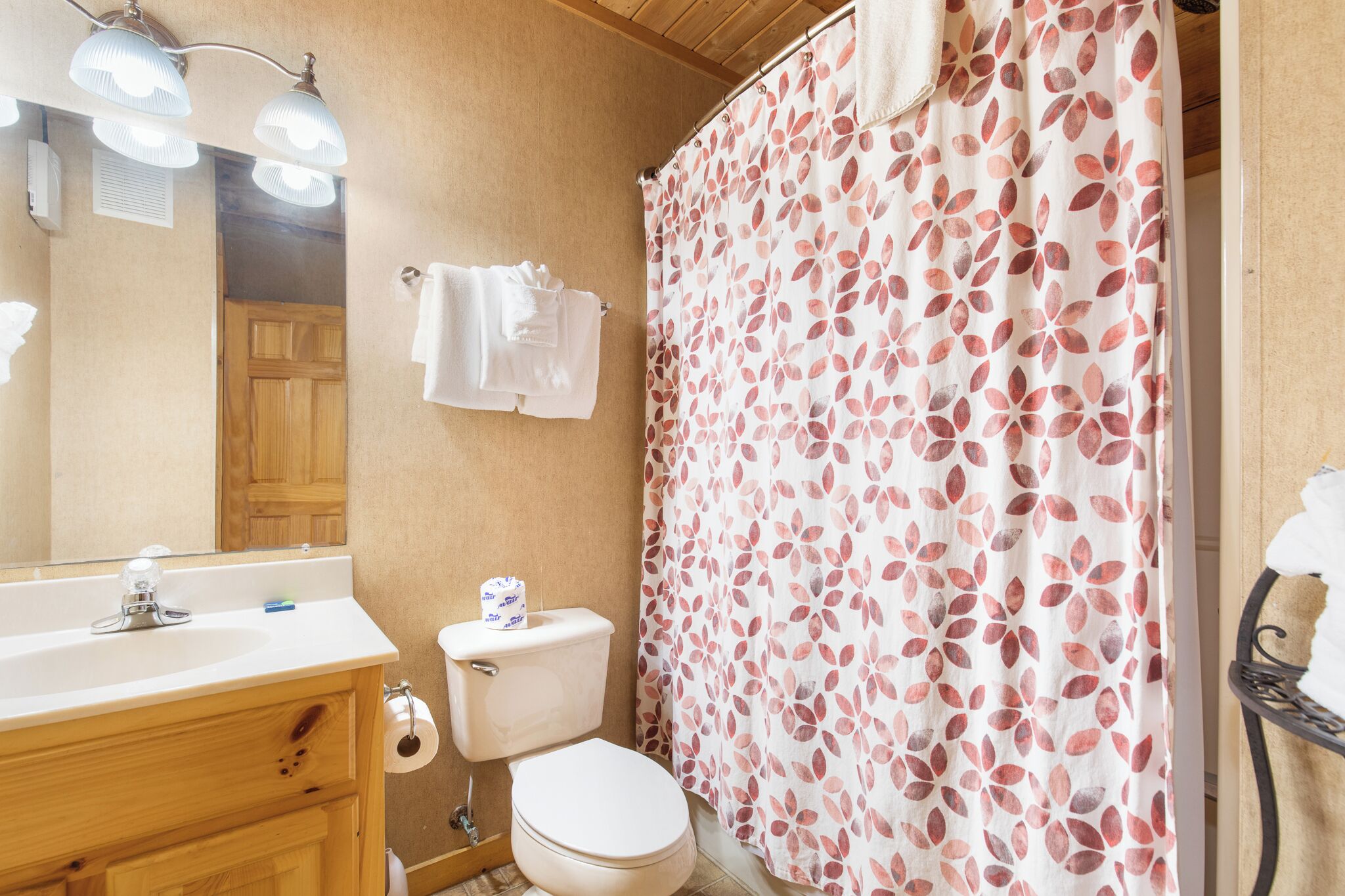 Un baño con tocador de madera, inodoro, cortina de ducha estampada con hojas y espejo con iluminación.