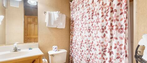 Un baño con tocador de madera, inodoro, cortina de ducha estampada con hojas y espejo con iluminación.