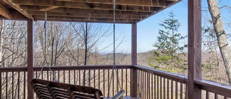 A wooden deck with a swing, surrounded by trees and a clear sky.