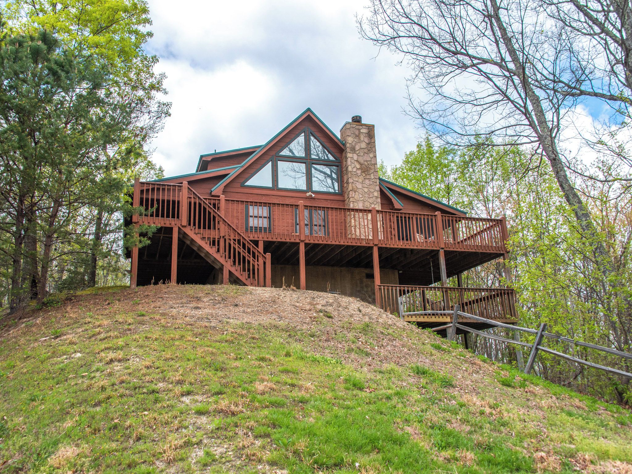 A two-story cabin with a stone chimney, wooden deck, and a staircase leading to a second level.
