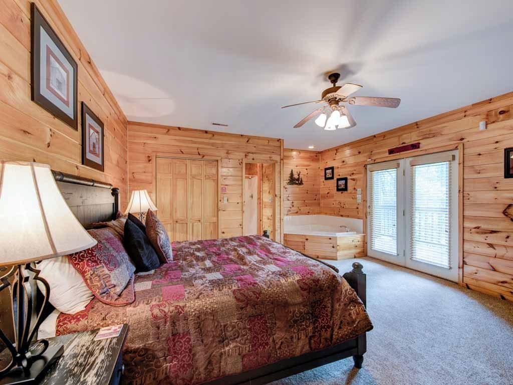 A wooden cabin bedroom with a bed, two bedside lamps, a ceiling fan, and a bathroom with a bathtub.