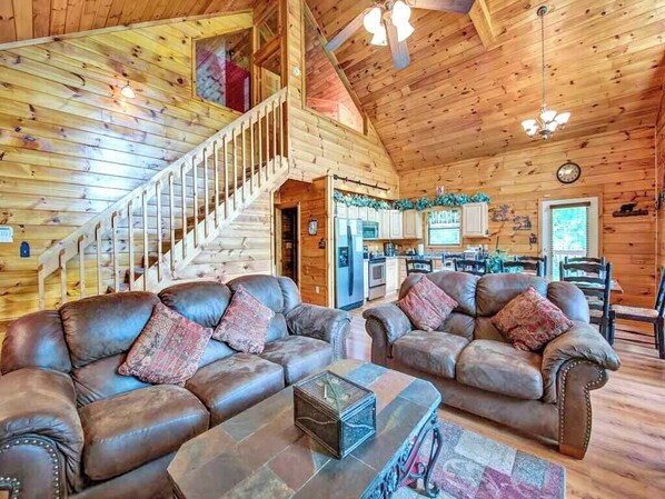 A spacious wooden cabin interior with a living area featuring two leather sofas, a stone coffee table, and a staircase leading to an upper level.