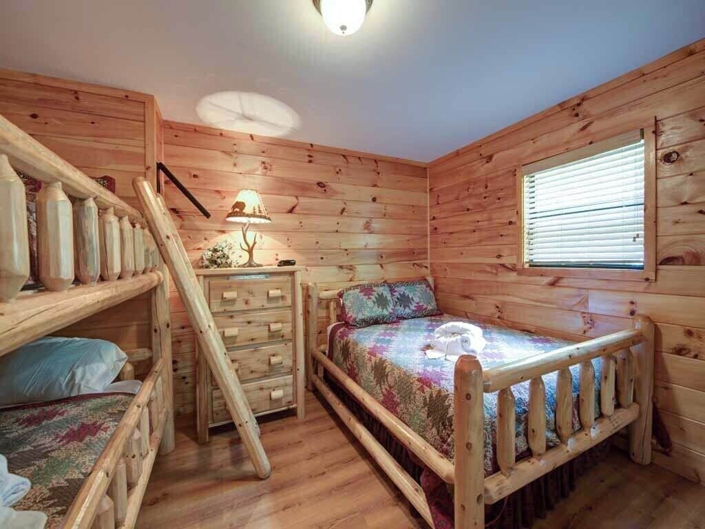 Une chambre dans une cabane en bois, avec des lits superposés, une commode, une lampe et une fenêtre avec des stores.