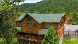 A log cabin with a balcony and a porch, surrounded by trees and a hilly landscape.