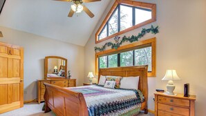A bedroom with a wooden bed, a nightstand, a dresser, a ceiling fan, and a large window with a view of trees. - Mountain Magic by Jackson Mountain Rentals (Sevierville)