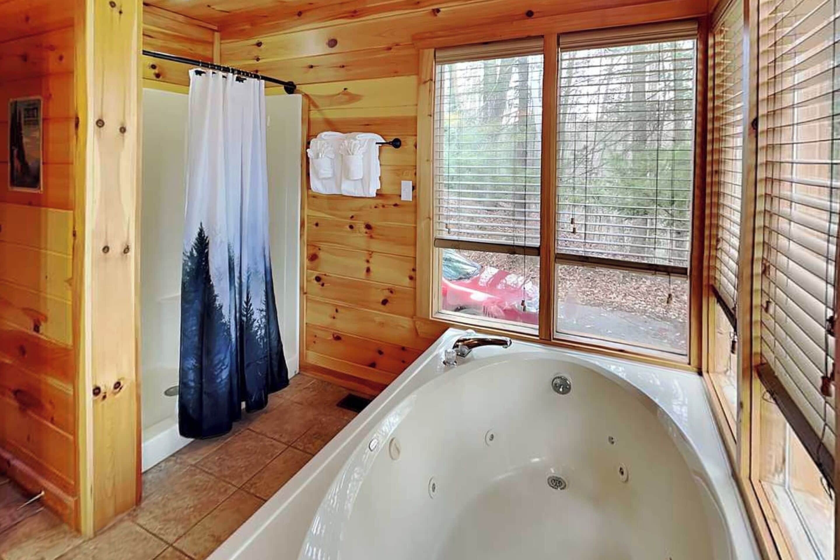 A wooden cabin bathroom with a large bathtub, a shower curtain with a forest print, and a window with blinds.