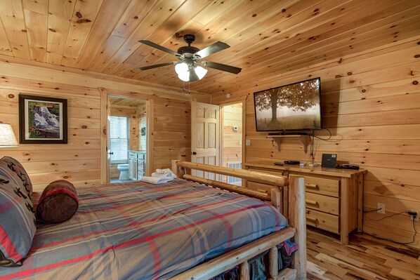 Quarto de cabana de madeira com cama, escrivaninha, televisão e ventoinha no teto.