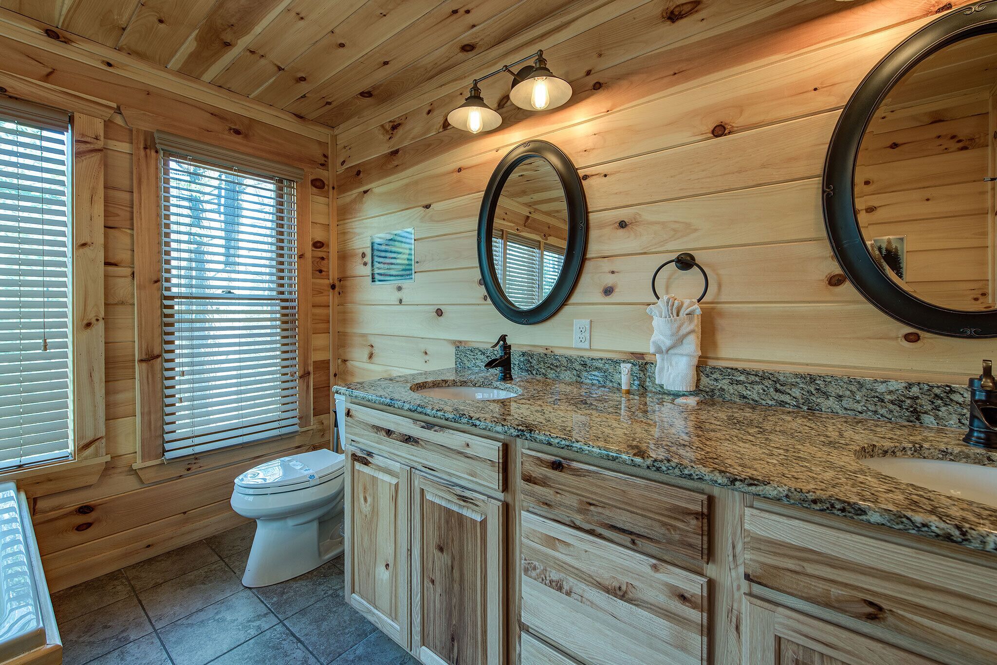 A wooden cabin bathroom with a granite countertop, two wall-mounted mirrors, a toilet, and a window with blinds.