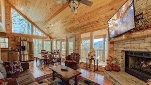 A spacious living room with a stone fireplace, wooden ceiling, and large windows offering a view of the outdoors. - Rocky Top Lookout by Jackson Mountain Rentals (Gatlinburg)