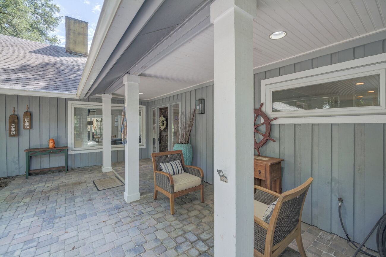 A covered outdoor area with a brick floor, wooden chairs, a bench, and a ship's wheel decoration.