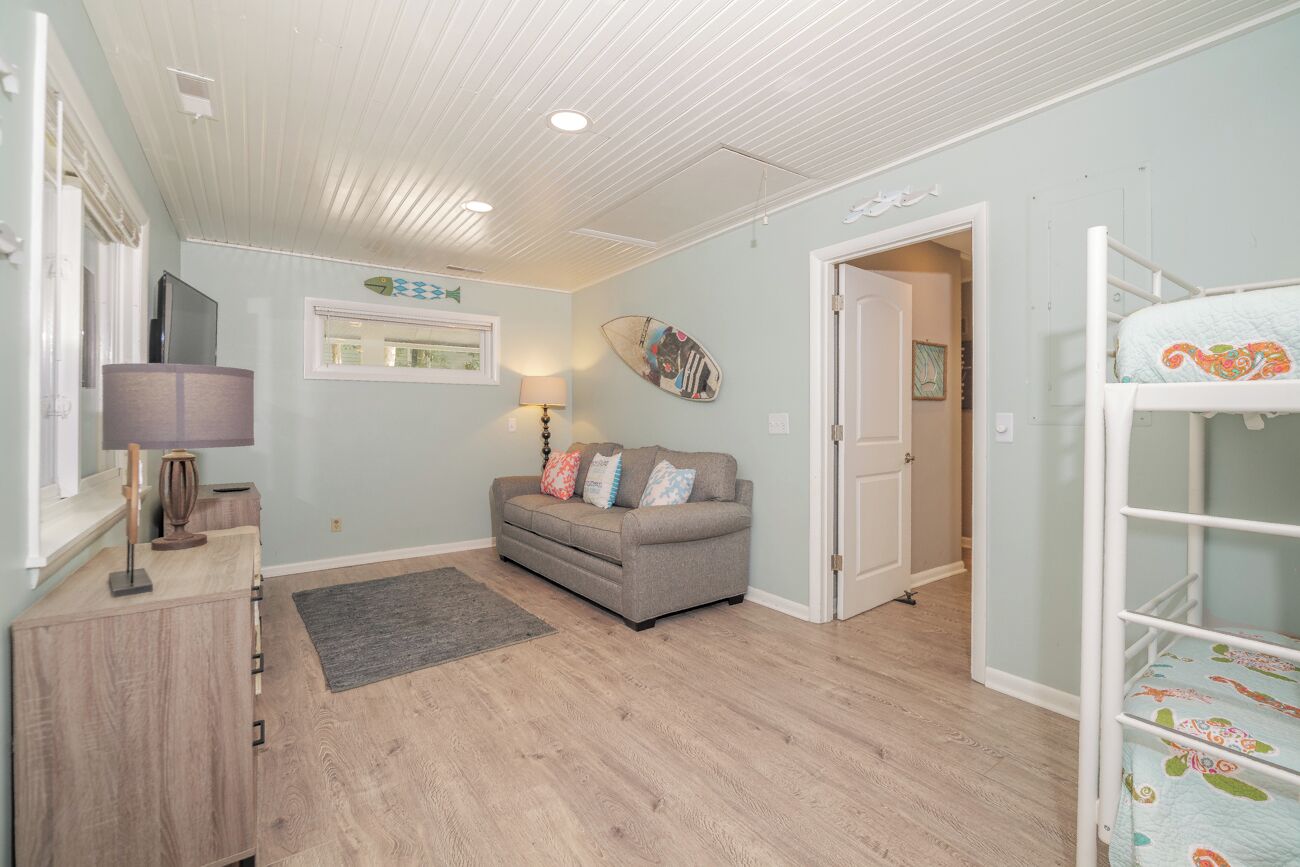 A room with a grey sofa, a wooden cabinet, a lamp, a surfboard, and a bunk bed.