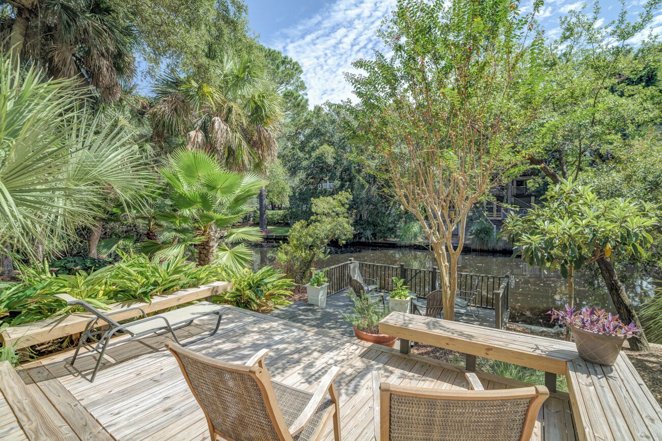 Une terrasse en bois, agrémentée de chaises et d’une table en osier, entourée d’une végétation luxuriante et d’un bassin.