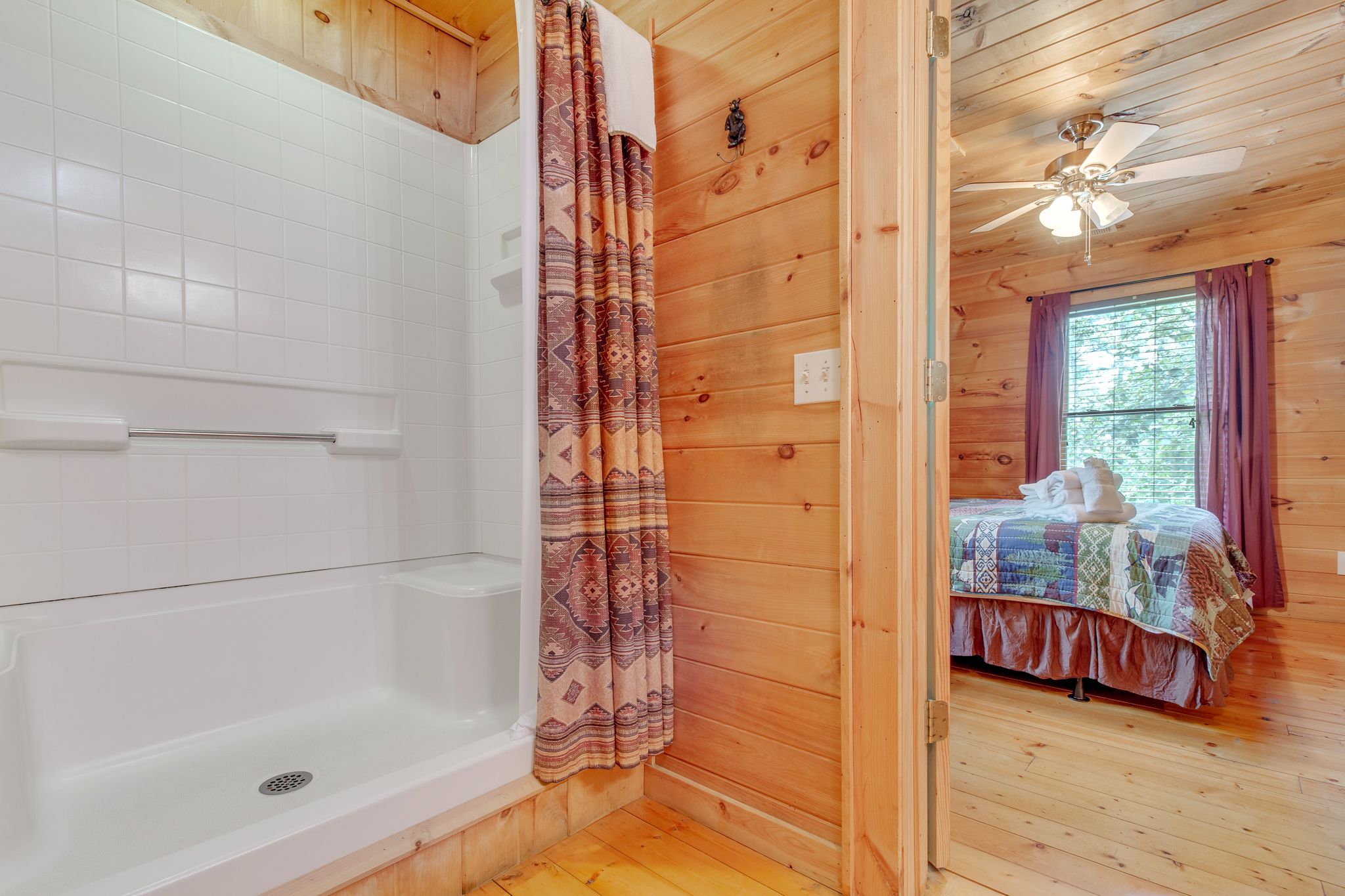A wooden cabin interior with a bathroom featuring a bathtub and a shower curtain, and a bedroom with a bed and a window.