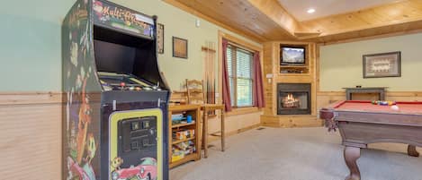 A room with a pool table, a multi-arcade machine, a television, and a fireplace.