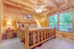 A wooden cabin bedroom with a bed, a ceiling fan, a window with blinds, and a small table with a lamp.