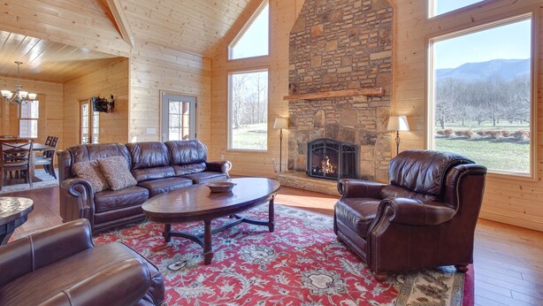 A log cabin interior with a stone fireplace, leather furniture, and large windows offering a view of the outdoors.