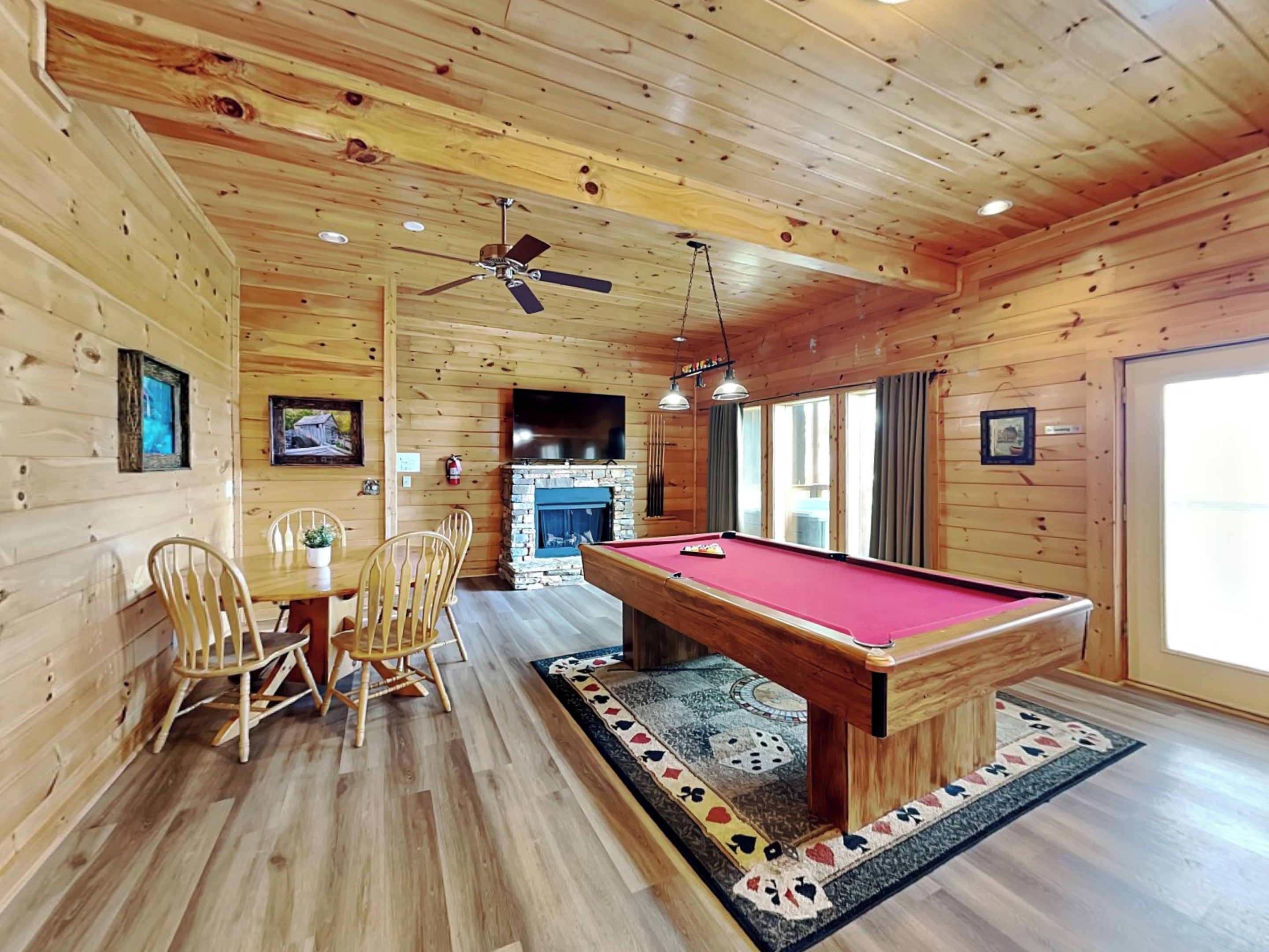 Interieur van een houten chalet met een pooltafel, stoelen, een open haard en een plafondventilator.