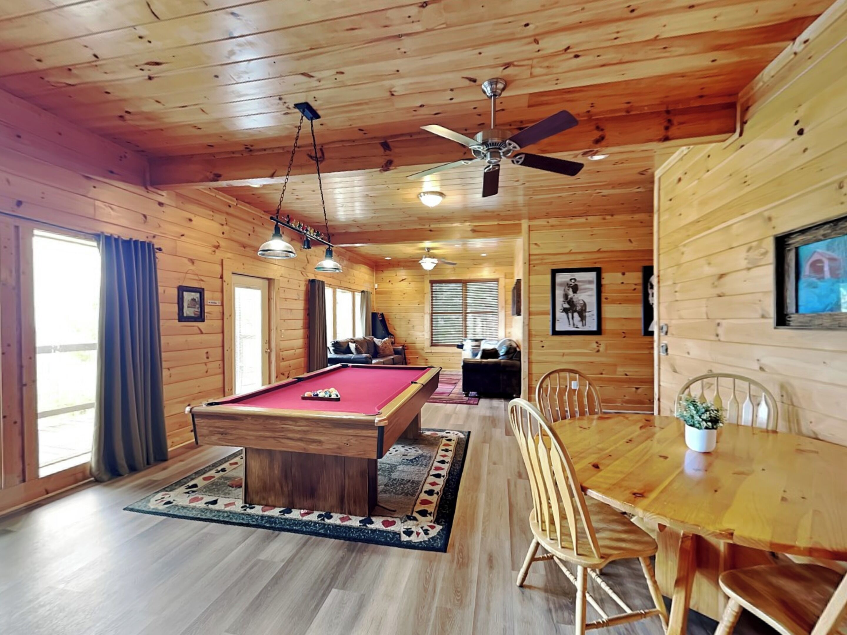 Interieur van een houten chalet met een pooltafel, een eettafel, stoelen, een plafondventilator en grote ramen.