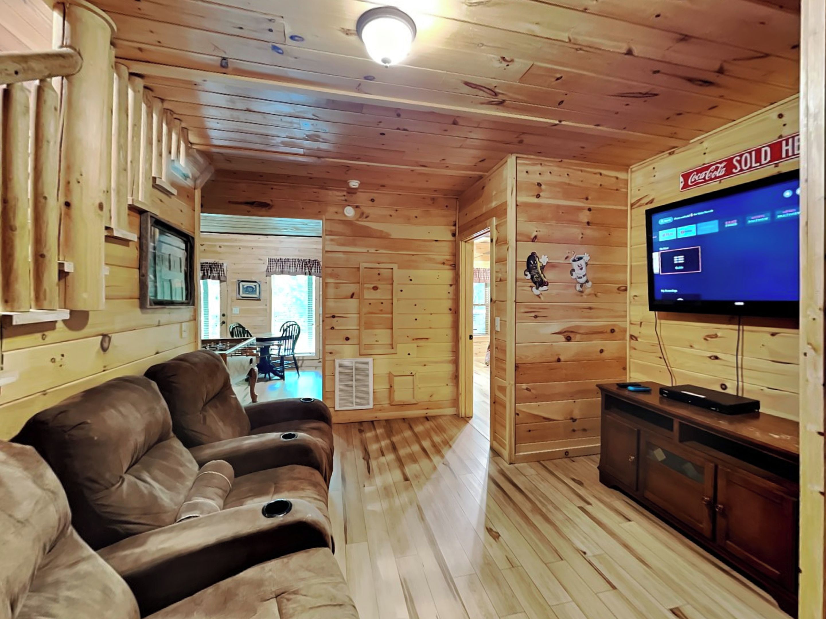 A cozy cabin interior with a wooden interior, a sofa, a TV, and a dining area visible through a doorway.
