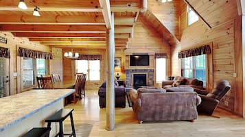 A log cabin interior with a dining area, a living room with a fireplace, and a large window.