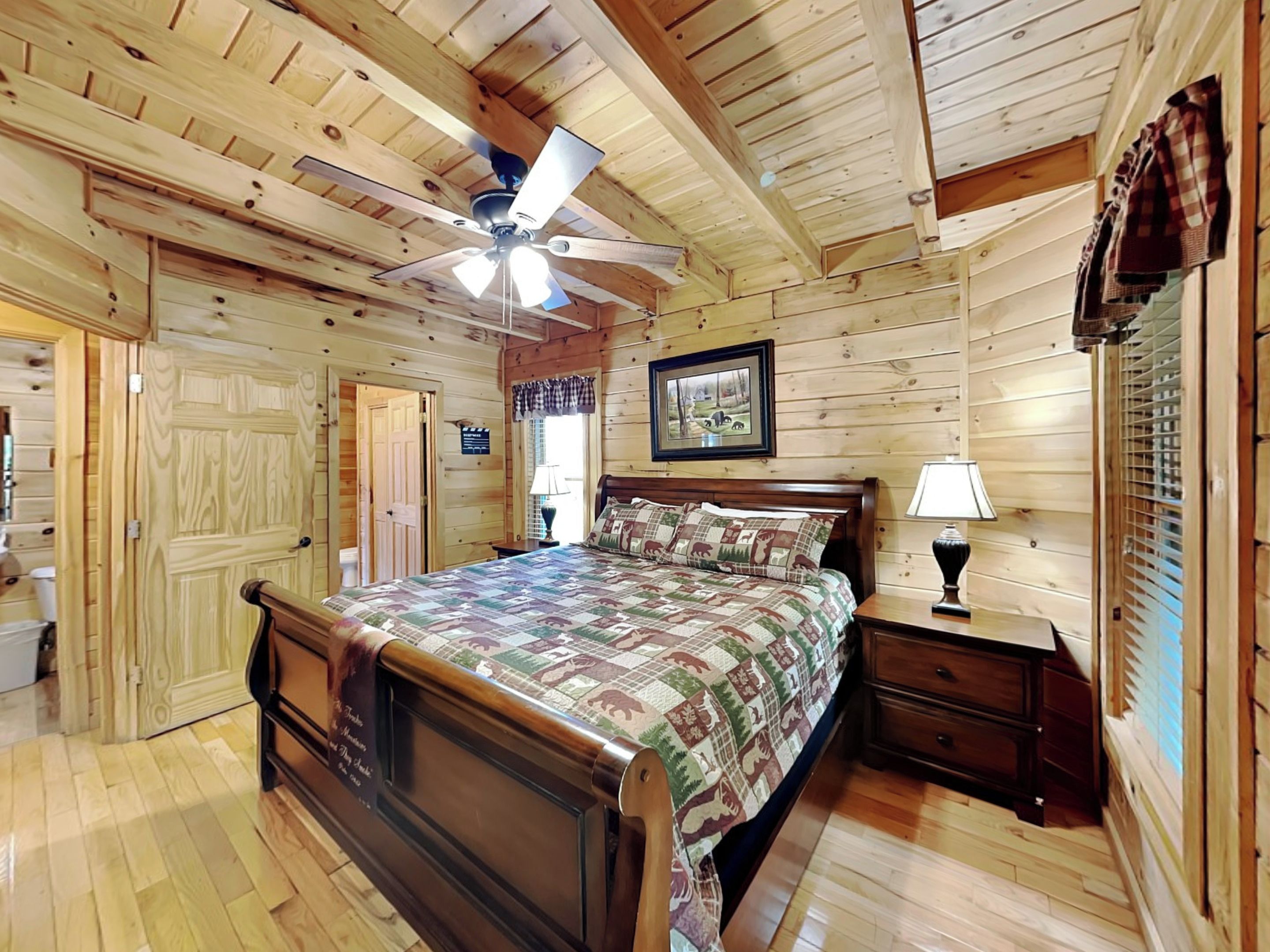 Un dormitorio de cabaña de madera con una cama, una mesita de noche, un ventilador de techo y una ventana con cortinas.
