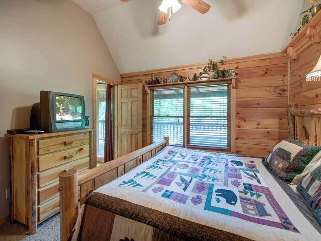 A bedroom with a wooden bed, a television on a wooden dresser, and a window with blinds.