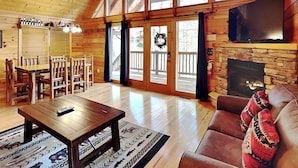 A log cabin interior with a fireplace, wooden furniture, and large windows. - Running Bear by Jackson Mountain Rentals (Gatlinburg)