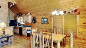 A rustic wooden interior with a dining area, kitchen, and a large window with blinds. - Running Bear by Jackson Mountain Rentals (Gatlinburg)