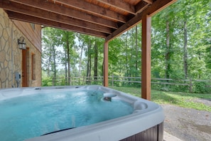 Un jacuzzi bajo una pérgola de madera, con un muro de piedra y una valla de madera en una zona boscosa.