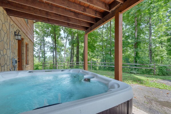 A hot tub under a wooden pergola with a stone wall and a wooden fence in a wooded area.
