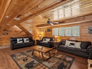 A cozy cabin interior with wooden walls, a ceiling fan, and two leather sofas with bear-themed cushions.