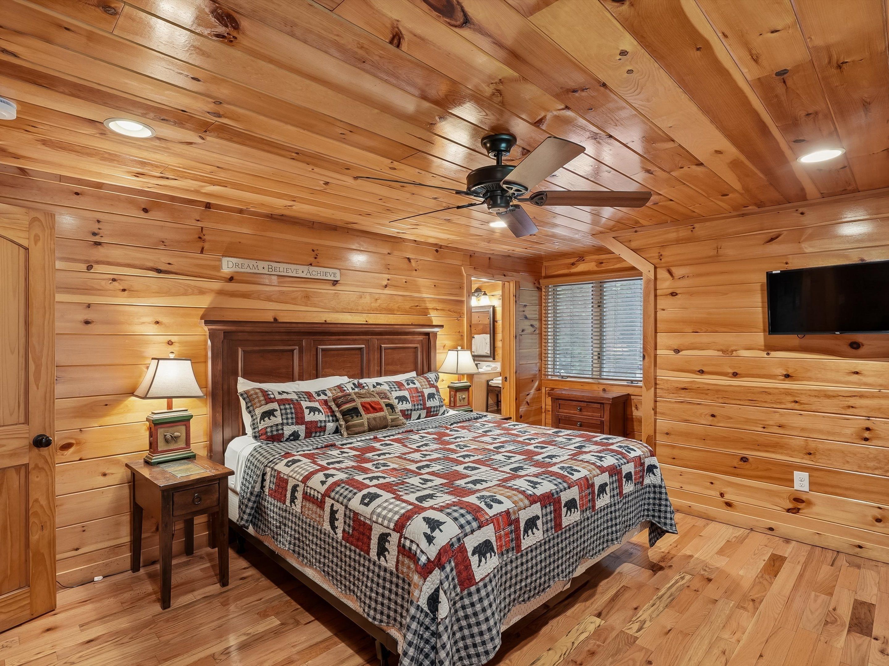 Une chambre de style cabane en bois, avec un lit, des tables de chevet, un ventilateur de plafond et un téléviseur fixé au mur.