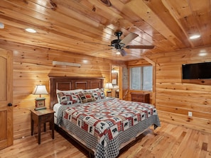 A wooden cabin bedroom with a bed, nightstands, a ceiling fan, and a television mounted on the wall. - Parkside Lodge by Jackson Mountain Rentals (Sevierville)