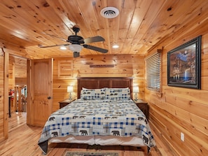 A wooden cabin bedroom with a bed, a ceiling fan, a nightstand, a framed picture, and a door leading to another room. - Parkside Lodge by Jackson Mountain Rentals (Sevierville)