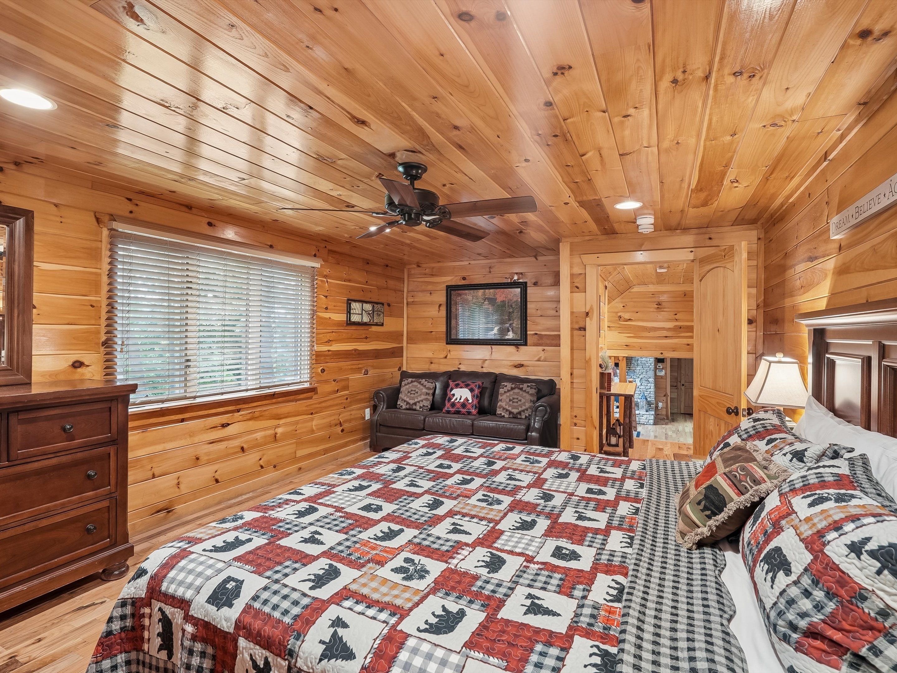 A log cabin room with a bed, a wooden dresser, a ceiling fan, and a window with blinds.
