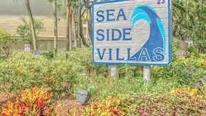 Opastekyltti, jossa lukee ”Sea Side Villas” ja jossa on numero ”23”. Kyltin ympärillä on vehreää kasvillisuutta ja värikkäitä kasveja.