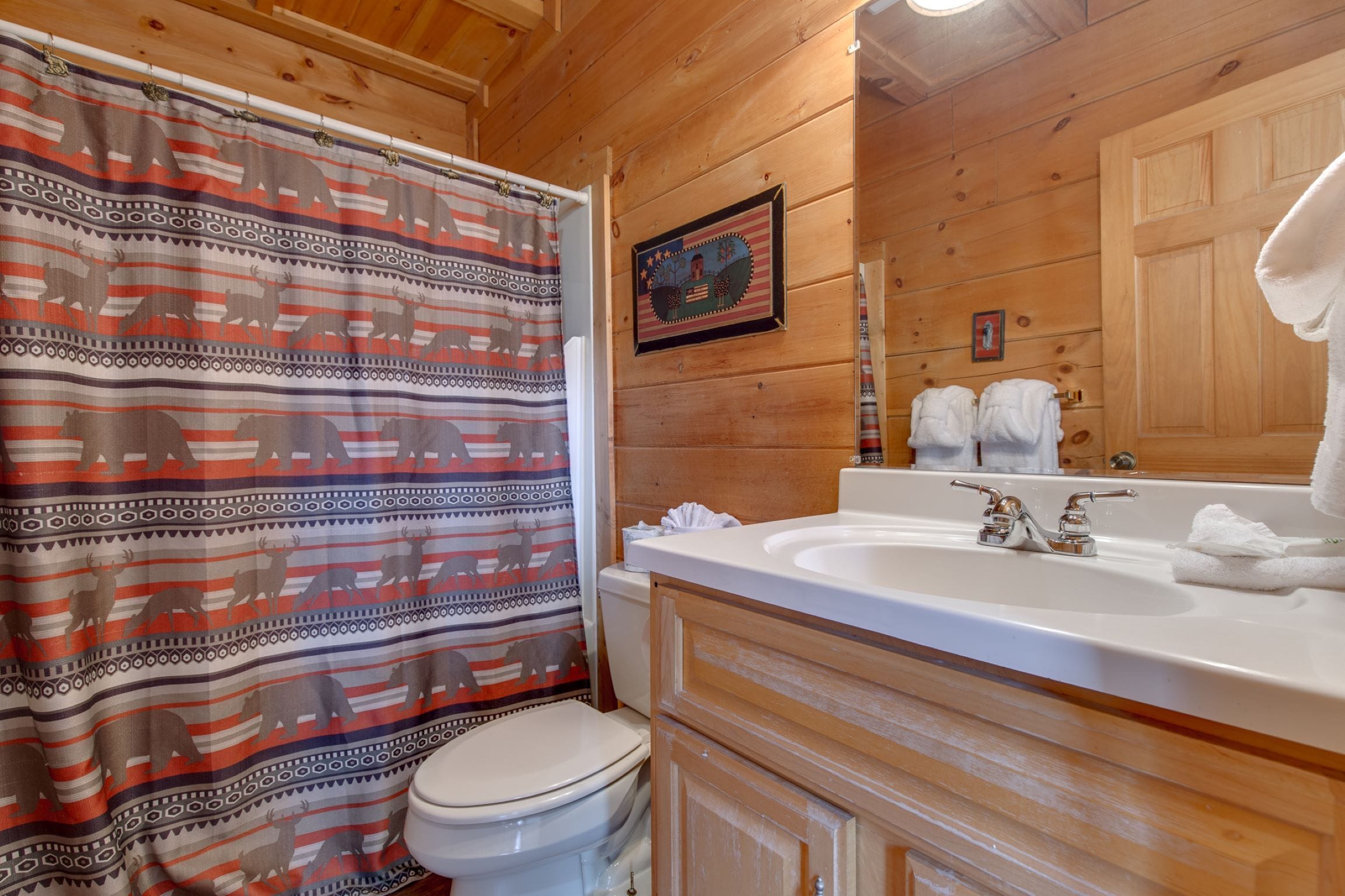 A wooden cabin bathroom with a shower curtain featuring animal prints, a white toilet, a sink with a mirror, and towels on the wall.