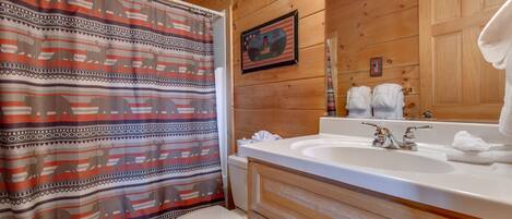 A wooden cabin bathroom with a shower curtain featuring animal prints, a white toilet, a sink with a mirror, and towels on the wall.