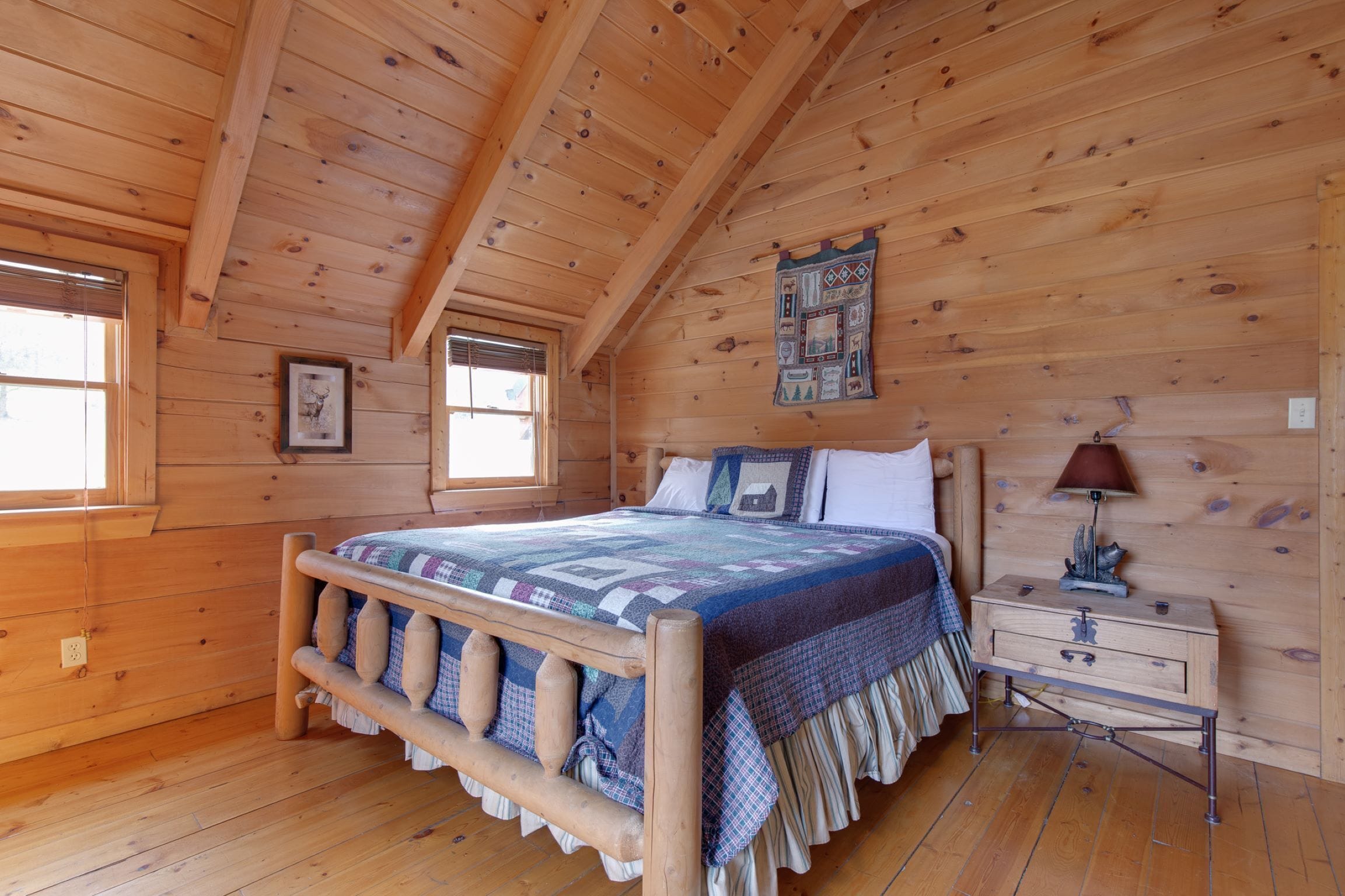 Un dormitorio de cabaña de madera con una cama, una mesita de noche, una lámpara y un tapiz acolchado en la pared.