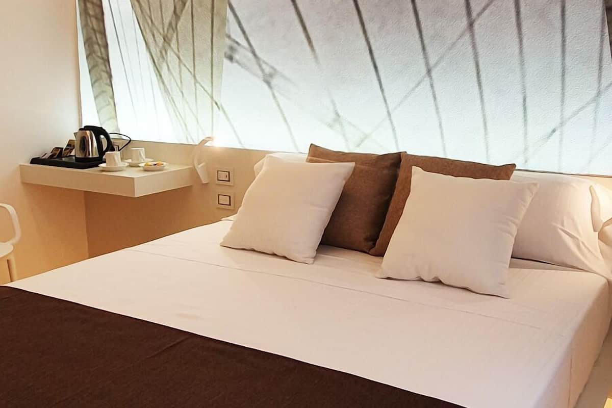 Quarto de hotel moderno com uma cama grande, mesa de cabeceira com telefone e cafeteira, e uma janela ampla com vista para uma ponte.