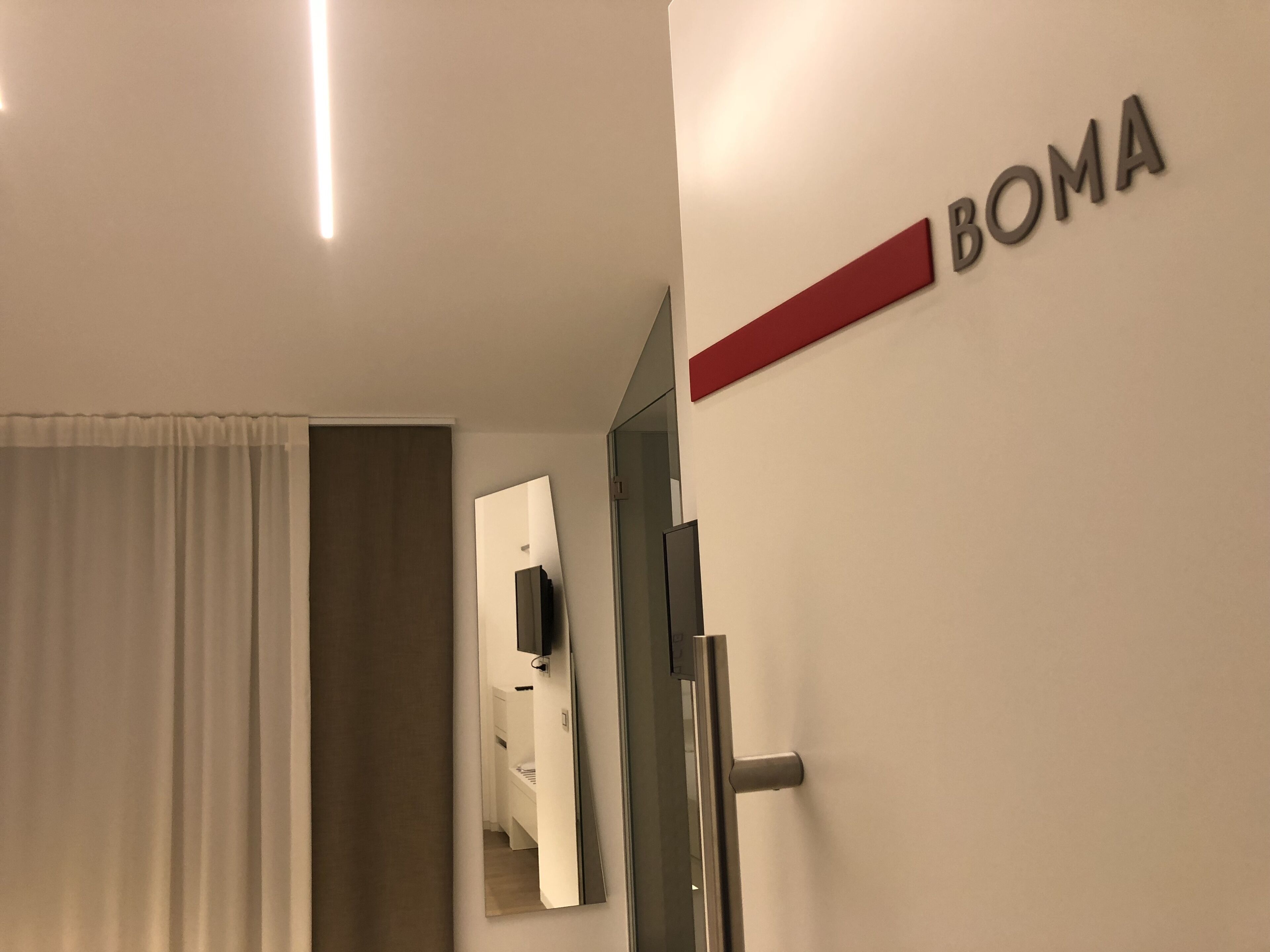Une chambre d’hôtel avec un lit, une porte, un miroir et une pancarte sur laquelle on peut lire « BOMA ».