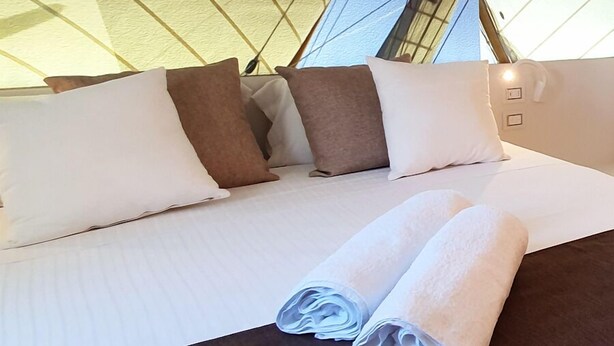 A bed with white and brown pillows, a neatly folded white towel, and a view of the building's exterior through a window. - B&B Gassa D’Amante (Vieste)